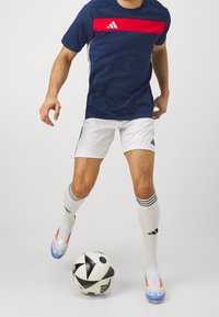Camiseta azul marino con una franja roja horizontal, pantalones cortos blancos, calcetines blancos con acentos negros y botas de fútbol con acentos azules, en posición para patear un balón de fútbol.
