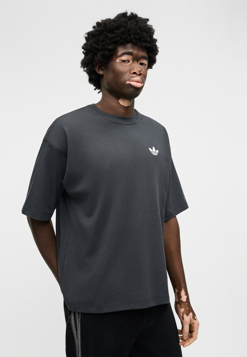 Junger Mann mit Vitiligo, der ein dunkelgraues Adidas-T-Shirt und schwarze Hosen trägt, vor einem schlichten hellen Hintergrund stehend.