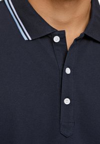 Polo shirt marine en tissu texturé, avec des boutons blancs et un col rayé bleu clair et blanc. Vue en gros plan du col.