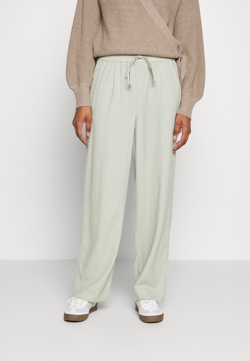 Personne portant un pull côtelé beige, un pantalon ample vert clair avec cordon et des baskets blanches, debout devant un fond uni.