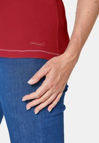 Rotes, tailliertes T-Shirt mit kurzen Ärmeln, glatter Textur und dezentem gestepptem Detail am Saum, kombiniert mit blauen Jeans.