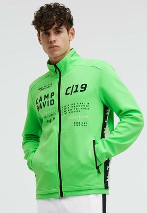 Young man wearing bright green zip-up jacket with black text and hands in pockets, against a plain light background.