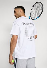 Balta marškiniai su juodu „Grand Slam Tour“ grafiniu vaizdu ant nugaros, derinami su pilkais šortais su šoninėmis juostomis. Laiko teniso raketę ir kamuoliuką.