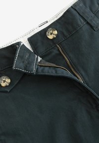 Pantalons vert foncé avec fermeture éclair et bouton, en tissu texturé avec des détails de couture visibles le long de la taille.
