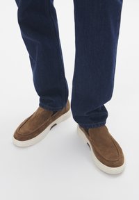 Hnědé semišové nazouvací boty s elastickými bočními panely, bílou gumovou podrážkou a hladkým povrchem, nošené s tmavě modrými džínami.