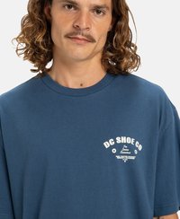 Man med lockigt axellångt hår och mustasch, iklädd en blå t-shirt från DC Shoe Co med vit text på bröstet.