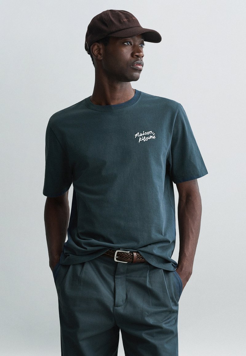 Dunkelblaues Baumwoll-T-Shirt mit weißem Schriftlogo, getragen mit schiefergrauen Hosen und einer braunen Mütze, das den lockeren Schnitt und die weiche Textur betont.