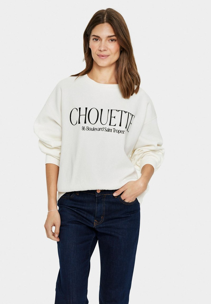 Fehér pulóver fekete felirattal: "CHOUETTE 86 Boulevard Saint Tropez", laza szabással és bordázott mandzsettával, sötét farmernadrággal párosítva.