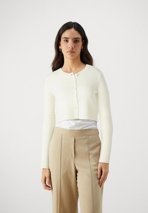 Cardigan court en tricot crème doux avec un col rond, des manches longues et une fermeture à boutons. Porté sur un haut blanc et un pantalon beige.