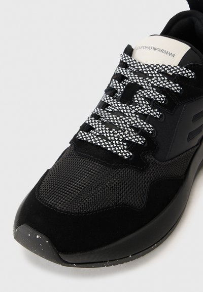 Black sneaker featuring a mesh and suede upper, textured black sole, bold branding on the tongue, and patterned black and white laces.