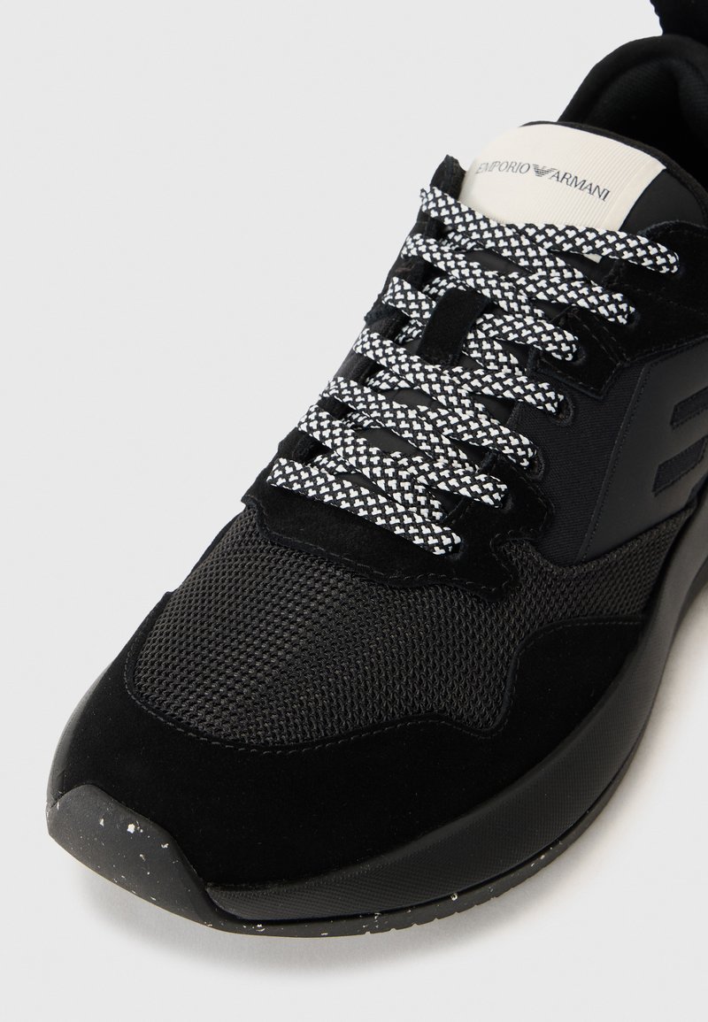 Zapatillas negras con un upper de malla y ante, suela texturizada negra, un atrevido logotipo en la lengüeta y cordones a rayas en blanco y negro.