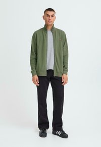 Grüne Reißverschlussjacke über einem grauen T-Shirt, kombiniert mit schwarzen Hosen und schwarzen Turnschuhen mit weißen Streifen. Einfaches Design, lässige Passform.