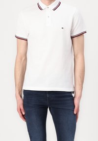 Polo blanc avec des accents de col et de manches rayés rouges et bleus, doté d'une patte de boutonnage à deux boutons et d'un petit logo sur la poitrine.