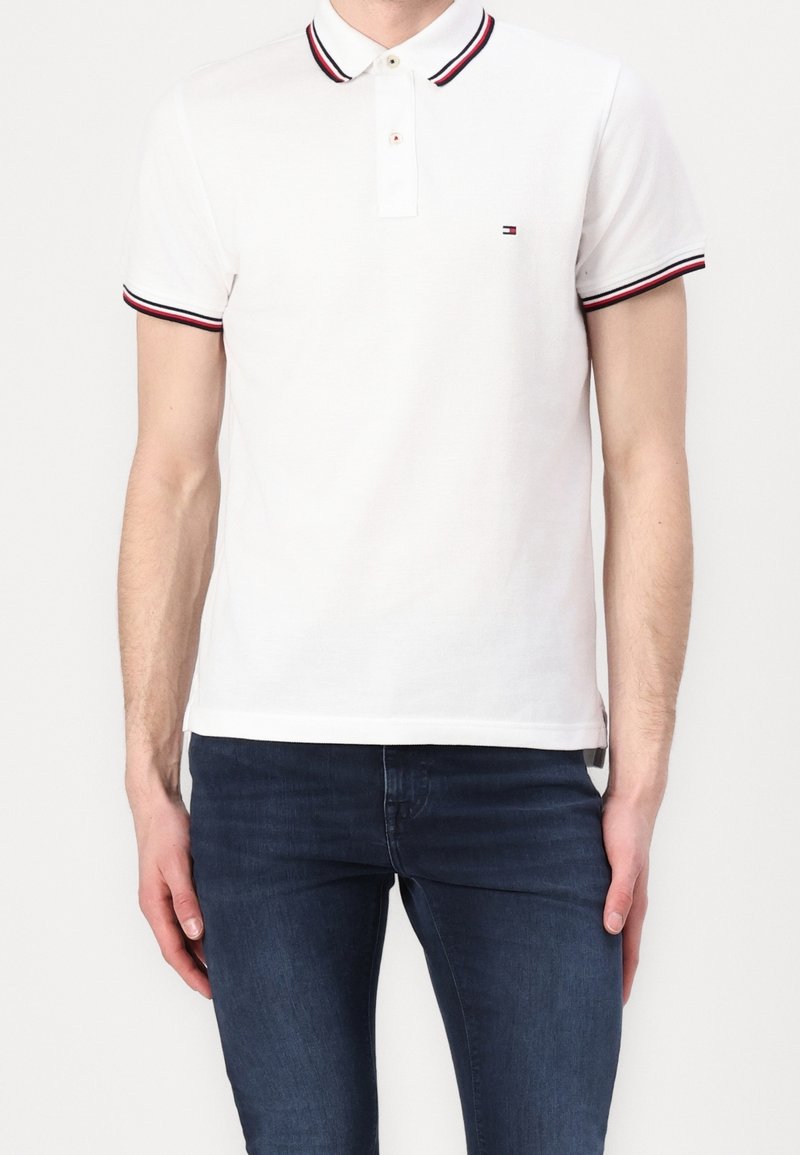 Polo blanc avec des accents de col et de manches rayés rouges et bleus, doté d'une patte de boutonnage à deux boutons et d'un petit logo sur la poitrine.