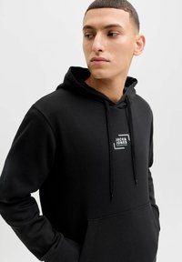 Sweat à capuche noir en mélange de coton, avec une poche avant, une capuche à cordon de serrage et une étiquette logo blanche sur la poitrine. Texture douce.