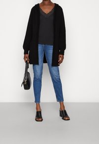 Cárdigan de punto negro con capucha, llevado sobre una camiseta de tirantes oscura con encaje, combinado con unos jeans ajustados de color azul claro y mulas negras de tacón cuadrado.