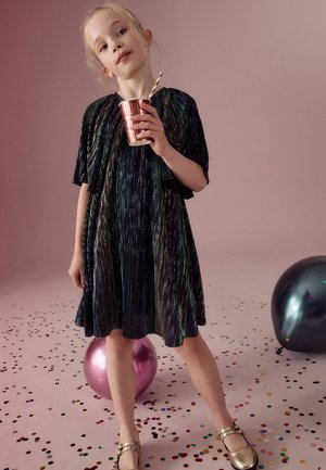 Robe noire et irisée à rayures avec une coupe ample et des manches courtes, accessoirisée avec une tasse en cuivre et des chaussures dorées, sur un fond rose.