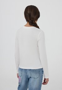 Camicia bianca a maniche lunghe con scollatura rotonda. Tessuto morbido, design aderente, abbinata a jeans in denim azzurro chiaro con dettagli grafici rosa.