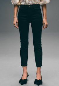 Jeans ajustados negros de cintura alta, con herrajes dorados, diseño clásico de cinco bolsillos y dobladillo a la altura del tobillo, combinados con tacones negros.