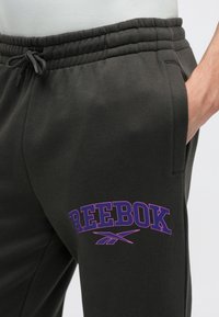 Tmavě šedé joggingové kalhoty z bavlněné směsi s elastickým pasem a fialovým logem 'Reebok' na levé straně. Disponují bočními kapsami a hladkým povrchem.