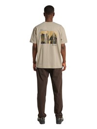 Mand iført beige T-shirt med by- og bjergtryk på ryggen, mørkebrune bukser og vandresko, stående med ryggen mod.