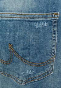 Zila denima auduma ar izbalinātām sekcijām, ar izšuvuma kabatas kontūru un dekoratīvu zelta izšuvumu. Nolietojuma detaļa netālu no kabatas.