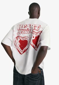 Tricou alb cu text grafic roșu "HEART struck" și două inimioare conturate pe spate, având un croi relaxat și mâneci scurte.