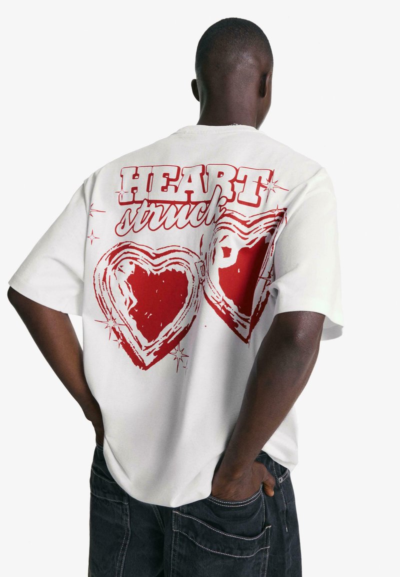 Tricou alb cu text grafic roșu "HEART struck" și două inimioare conturate pe spate, având un croi relaxat și mâneci scurte.