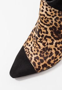 Botín de tobillo de punta afilada con punta de ante negro y parte superior con estampado de leopardo sobre un fondo blanco.
