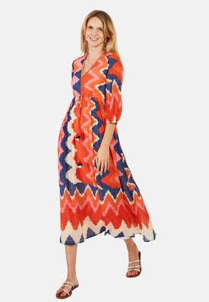 Woman in a long, colorful zigzag-patterned dress with half sleeves, smiling and wearing strappy sandals on a plain light background.