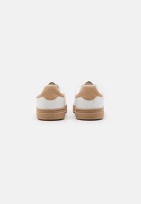 Białe sneakersy z brązowymi zamszowymi akcentami, okrągłym obcasem i gumową podeszewką. Posiadają małe logo na obcasie. Gładka textura z czystymi liniami.