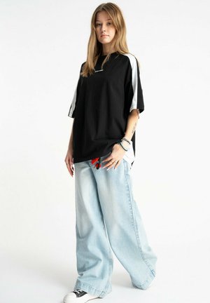 Młoda kobieta stojąca, ubrana w oversize'owy czarno-biały T-shirt, szerokie jasnoniebieskie dżinsy z szerokimi nogawkami, czarno-białe sneakersy oraz bransoletki.