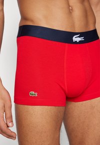 Boxer briefs de algodão vermelhos com uma cinta azul-marinho, apresentando um pequeno logótipo de crocodilo bordado em verde no lado esquerdo. Textura suave.