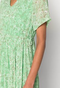 Blommig klänning i ljusgrön tyg med v-ringning, korta ärmar och samlad midja. Har ett strukturerat blommönster och knytdetalj.