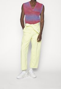 Hombre vestido con un suéter sin mangas de degradado rojo y púrpura con cuello en V, pantalones de cintura alta de color amarillo pálido con un cinturón atado y zapatillas blancas.