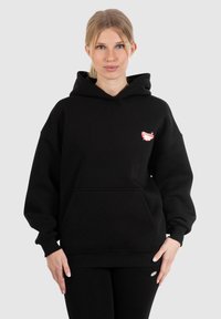 Sweatshirt à capuche noir en tissu doux. Doté d'une poche frontale et d'un design coloré brodé sur la poitrine gauche. Coupe régulière.