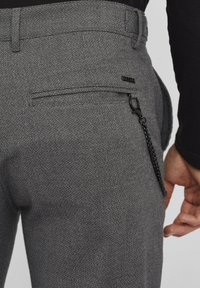Gros plan sur une personne portant un pantalon gris texturé avec une poche arrière, une petite étiquette de marque noire et une chaîne noire attachée.