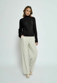 Schwarzer gerippter Rollkragenpullover kombiniert mit cremfarbenen weit geschnittenen Hosen. Das Model steht mit den Händen in den Taschen und trägt hellfarbene Schuhe.