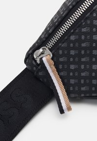 Černá taška s texturovaným povrchem, který má geometrický vzor, stříbrný zip a popruh s hnědým a bílým akcentem.