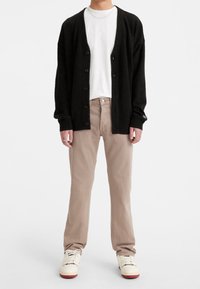 Cardigan negro de punto con botones, camiseta blanca, chinos beige y zapatillas crema con suelas rojas, de pie sobre un fondo liso.