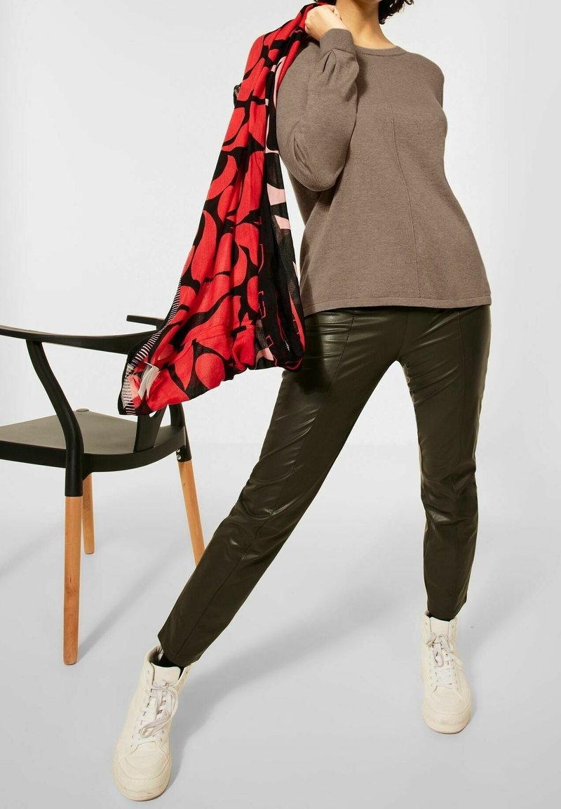 Personne portant un pull marron, un pantalon en cuir noir, des baskets blanches, tenant une écharpe rouge et noire à motifs à côté d'une chaise noire aux pieds en bois.