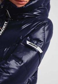 Mørkeblå pufferjakke med en glat og skinnende overflade. Har en høj krave og et fremtrædende "JACK1T"-mærke på ærmet. Kviltet design er synligt.