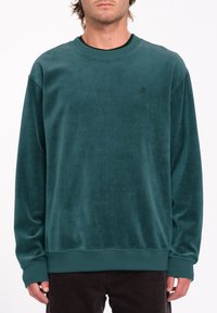 Sudadera de terciopelo verde azulado con cuello redondo, puños y dobladillo acanalados, y un sutil detalle del logo en el pecho. Textura suave y corte relajado.