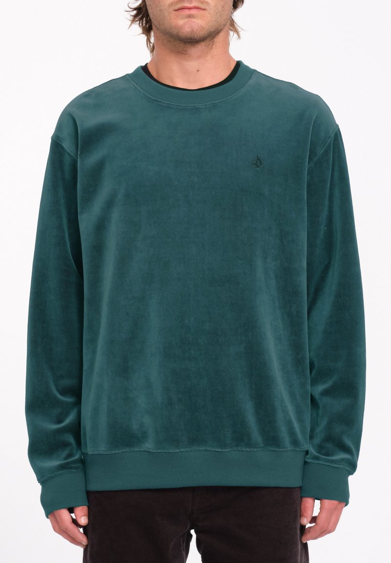 Sudadera de terciopelo verde azulado con cuello redondo, puños y dobladillo acanalados, y un sutil detalle del logo en el pecho. Textura suave y corte relajado.