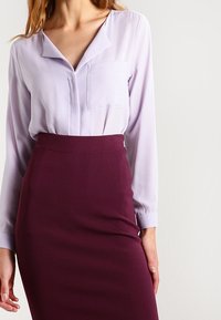 Blusa lila claro con escote en V y un solo bolsillo en el pecho, combinada con una falda lápiz ajustada de burdeos oscuro. Textura de tela suave, mangas largas.