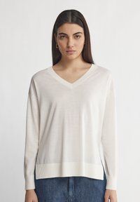 V-Ausschnitt, langärmeliger Pullover in hellem Beige, aus weichem Stoff gefertigt. Mit geripptem Ausschnitt und Bündchen, lockerem Schnitt und seitlichen Schlitzen am Saum.
