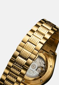 Goldene Uhr mit einem gemusterten Metallarmband, das geometrische Designs aufweist. Die Rückseite zeigt komplizierte Maschinendetails und ein rundes Gehäuse.