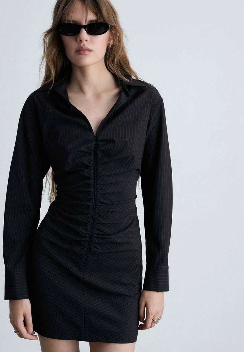 Rochie neagră cu fermoar, cu mâneci lungi, cu dungi verticale și texturi pliate pe torace, asortată cu ochelari de soare negri.