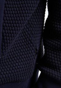 Marinblått strukturerat tyg med ett bikake-mönster, som har en slät panel och en liten logotagg, vilket visar en blandning av stickade stilar.