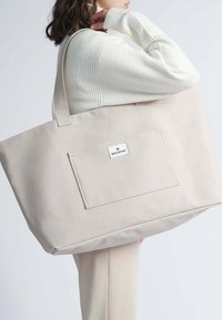 Bolsa tote de lienzo beige con dos asas resistentes y un bolsillo frontal. Presenta una etiqueta de marca en el bolsillo y una textura suave y duradera.